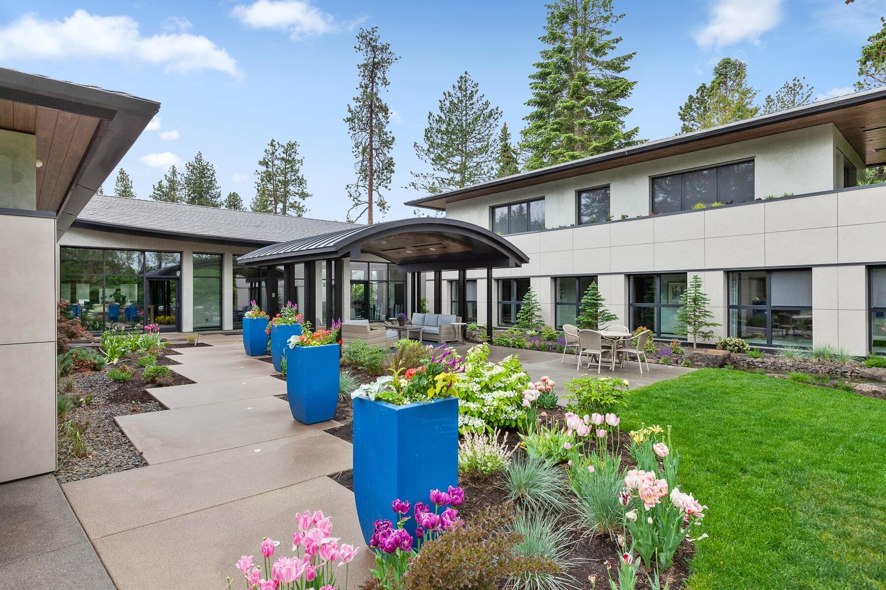 Modern two-story building with large windows, courtyard, blue planters, flowers, and outdoor seating.