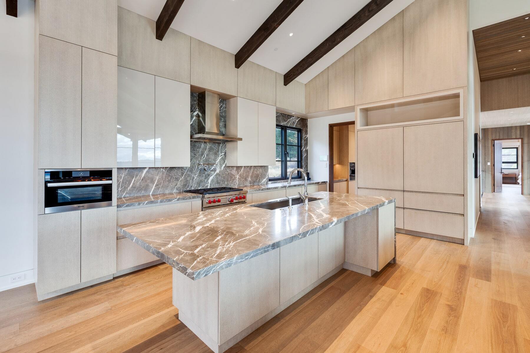 Modern kitchen with light wood cabinets, large island, marble backsplash, exposed beams, and windows.