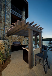 Outdoor stone kitchen with pergola and lake view.