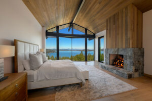 Modern bedroom with lake view, stone fireplace, wood ceiling, and large windows. Cozy and serene atmosphere.